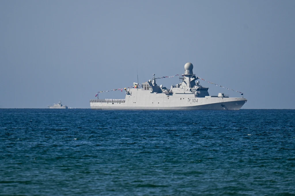 Wojenny okręt nawodny z rozwiniętymi flagami płynie po spokojnym morzu, na horyzoncie widoczna jest mniejsza jednostka.