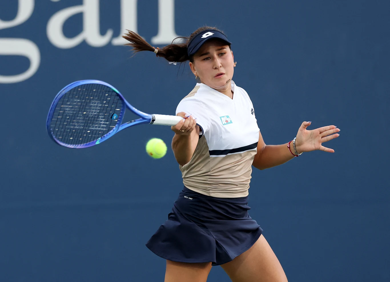 Tenisistka w sportowym stroju podczas gry, skoncentrowana na odbiciu żółtej piłki tenisowej rakietą, w tle gładka, niebieska ściana z fragmentem napisu.