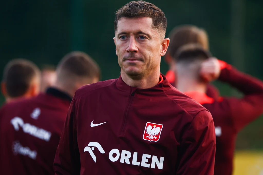 Robert Lewandowski Mężczyzna w bordowym dresie z logo PZPN i Orlen, stojący na tle kilku innych osób przygotowujących się do treningu sportowego. Twarz skupiona, światło naturalne, tło rozmazane wskazujące na aktywność sportową.