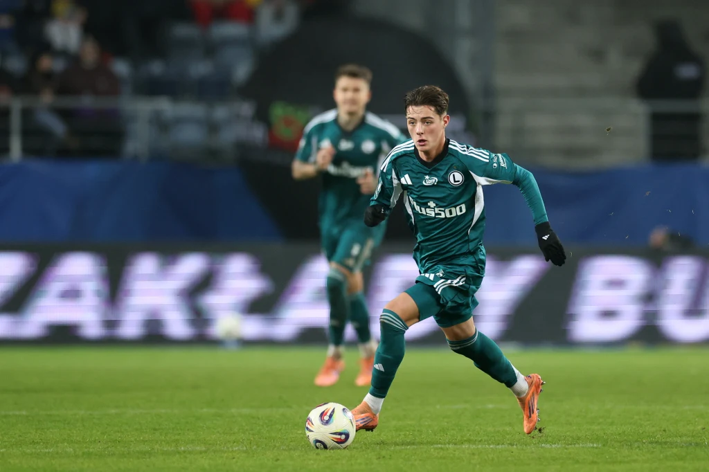 Kacper Urbański w barwach Legii Warszawa Piłkarz w zielonym stroju biegnący z piłką po murawie podczas meczu piłki nożnej, w tle drugi zawodnik tej samej drużyny oraz widownia.