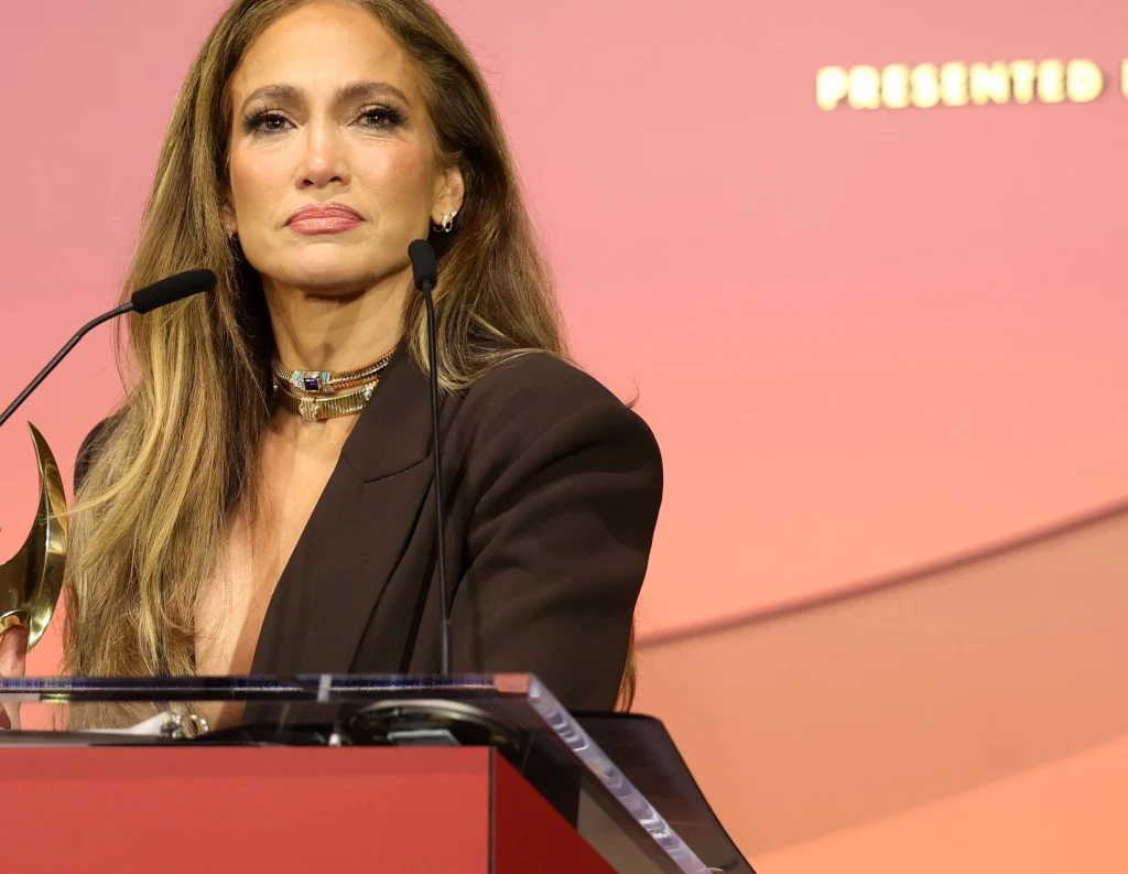 Jennifer Lopez podczas gali Women in Entertainment Kobieta o długich, jasnych włosach stoi za mównicą z mikrofonami, ubrana elegancko w ciemną marynarkę, na szyi ma złoty naszyjnik, w tle jasnoczerwone tło z elementami tekstu.