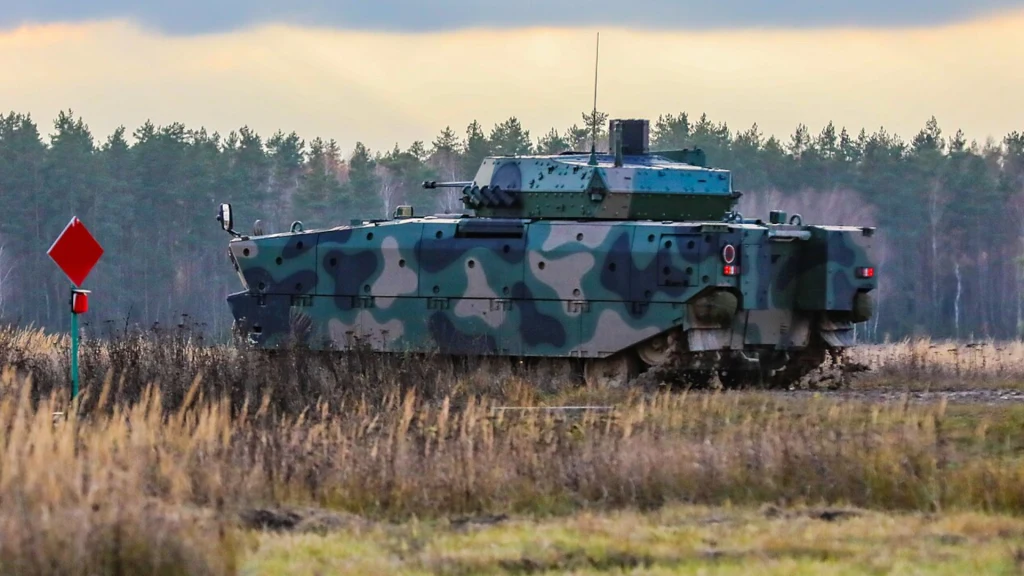 Pojazd opancerzony w kamuflażu leśnym jedzie po poligonie, otoczony wysoką suchą trawą oraz sosnowym lasem w tle, widoczny jest także czerwony znak drogowy po lewej stronie.