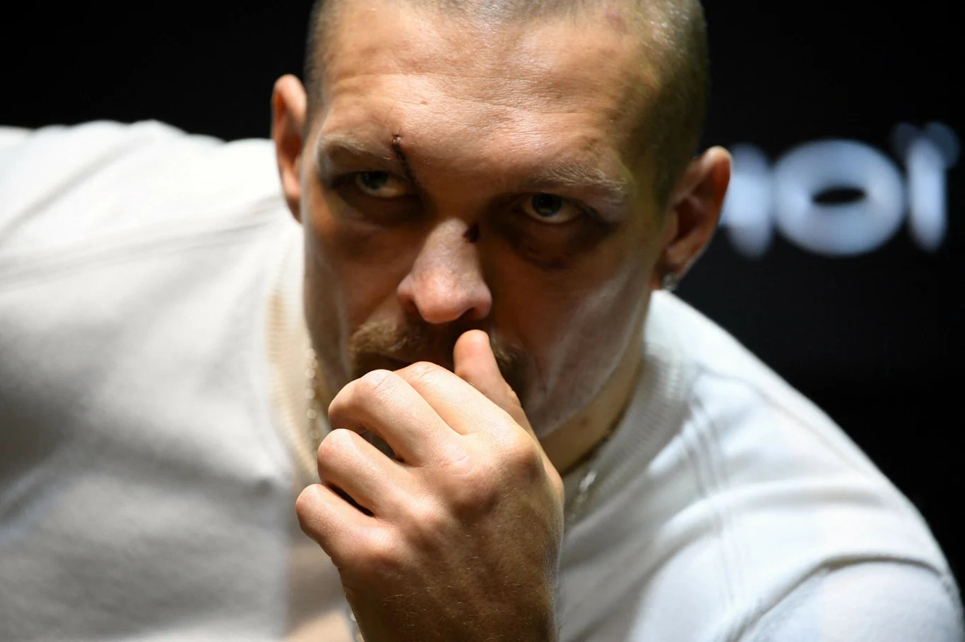 Ołeksandr Usyk Mężczyzna z podbitym okiem i rozcięciem nad brwią w białej koszulce, skupiony i zamyślony, opiera dłoń na ustach, na czarnym tle.