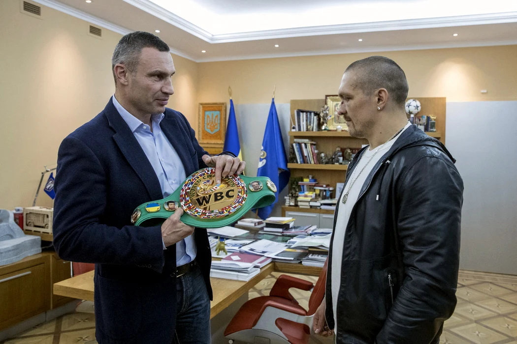 Witalij Kliczko oraz Ołeksandr Usyk podczas spotkania w Kijowie przed czterema laty Dwóch mężczyzn rozmawia w biurze, jeden z nich trzyma pas mistrzowski WBC. W tle widoczne są ukraińskie flagi, dokumenty na biurku i regał z książkami.
