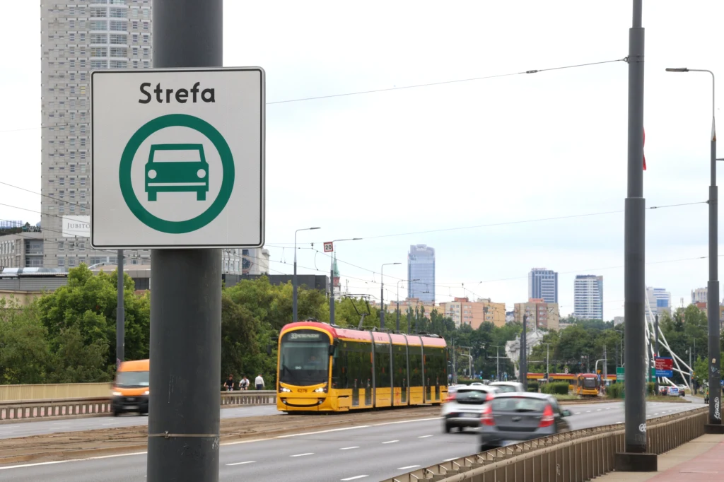 Jasnożółty tramwaj jedzie po wydzielonym torowisku obok ruchliwej ulicy, na której poruszają się samochody. Na pierwszym planie widoczny znak informujący o strefie tranzytowej dla samochodów. W tle nowoczesne wieżowce i zielone drzewa.