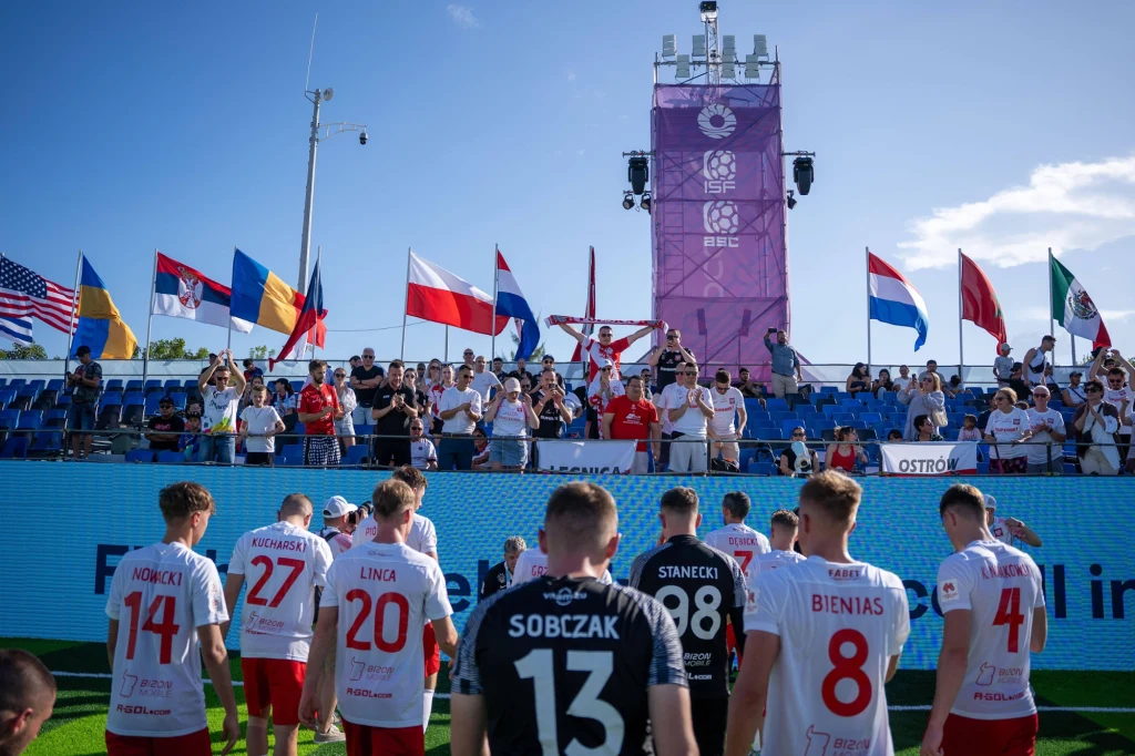 Reprezentacja Polski w futbolu sześcioosobowym Drużyna piłkarska ubrana w białe i czerwone stroje stoi na boisku zwrócona plecami do aparatu, przed trybunami pełnymi kibiców, nad którymi powiewają flagi wielu krajów, w tym Polski. W tle widoczne są konstrukcje i telebimy związane z organizacją wyda...