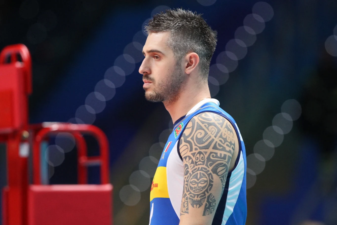 Siatkarz w sportowym stroju z widocznym dużym tatuażem na ramieniu stoi na tle halowej areny, w tle rozmazane światła ozdobne i element czerwonej struktury.