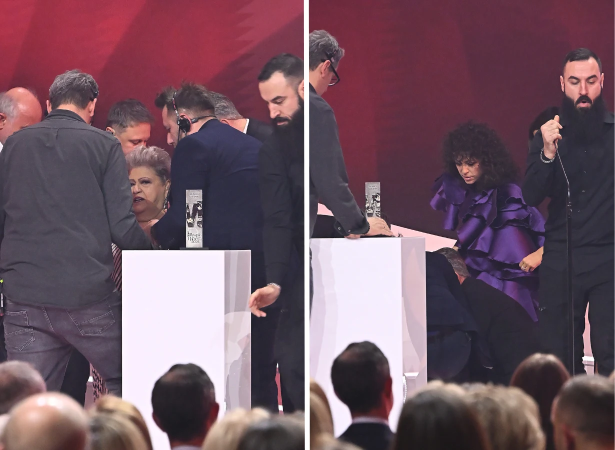 Grupa osób zgromadzona wokół podium podczas oficjalnej gali. Na pierwszym planie widoczne są dwie kobiety stojące i siedzące przy białym postumencie, w tle kilku mężczyzn, a całość odbywa się na tle czerwonej scenografii.