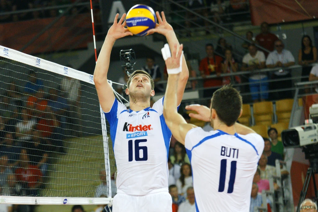 Michele Baranowicz w reprezentacji Włoch Dwóch siatkarzy w biało-niebieskich strojach rozgrywa piłkę nad siatką w hali sportowej, dookoła widoczna publiczność oraz sprzęt telewizyjny.