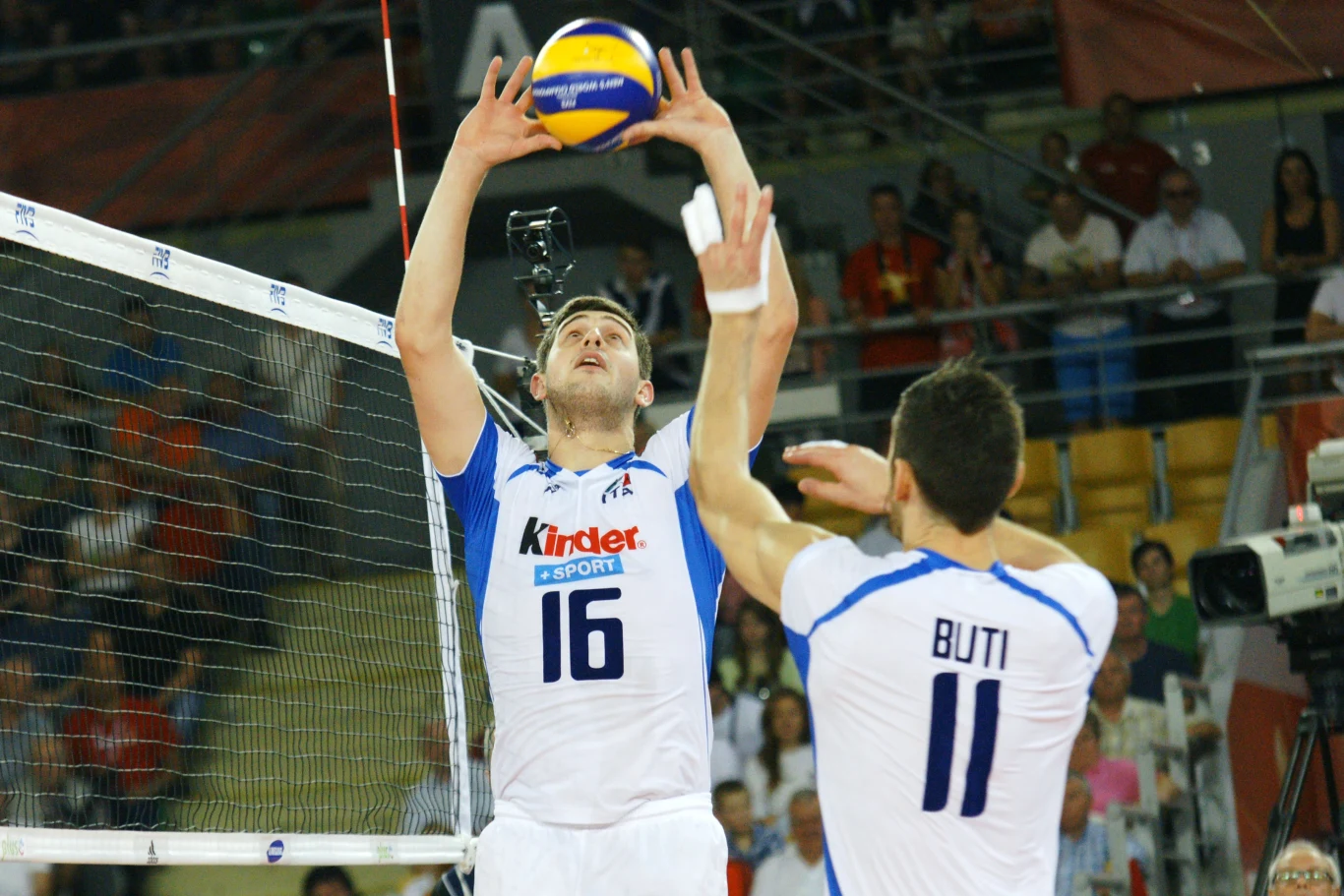 Dwóch siatkarzy w biało-niebieskich strojach rozgrywa piłkę nad siatką w hali sportowej, dookoła widoczna publiczność oraz sprzęt telewizyjny.