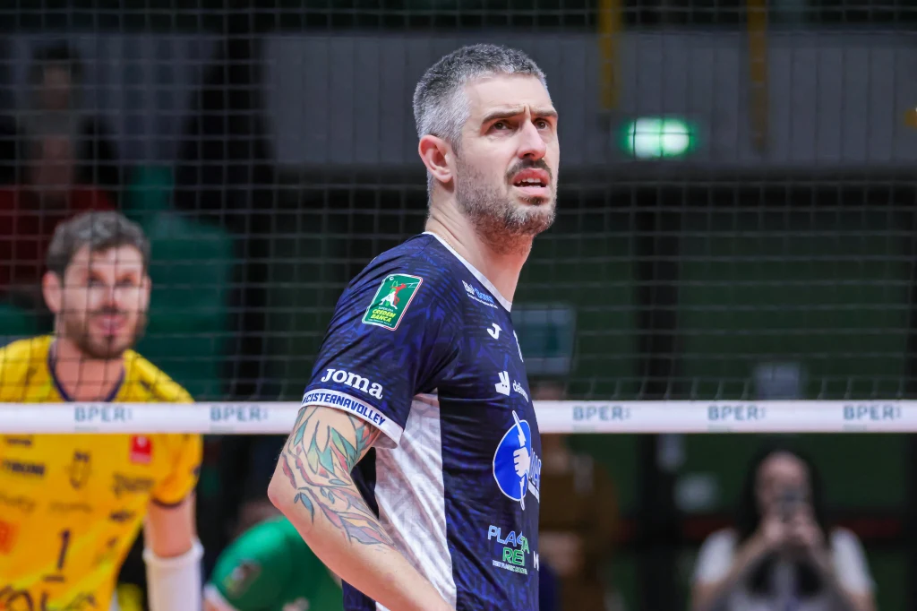 Michele Baranowicz Siatkarz w ciemnej koszulce z wyraźnym tatuażem na ramieniu stoi przy siatce na hali sportowej, w tle widoczny zawodnik w żółtym stroju oraz fragmenty trybun.