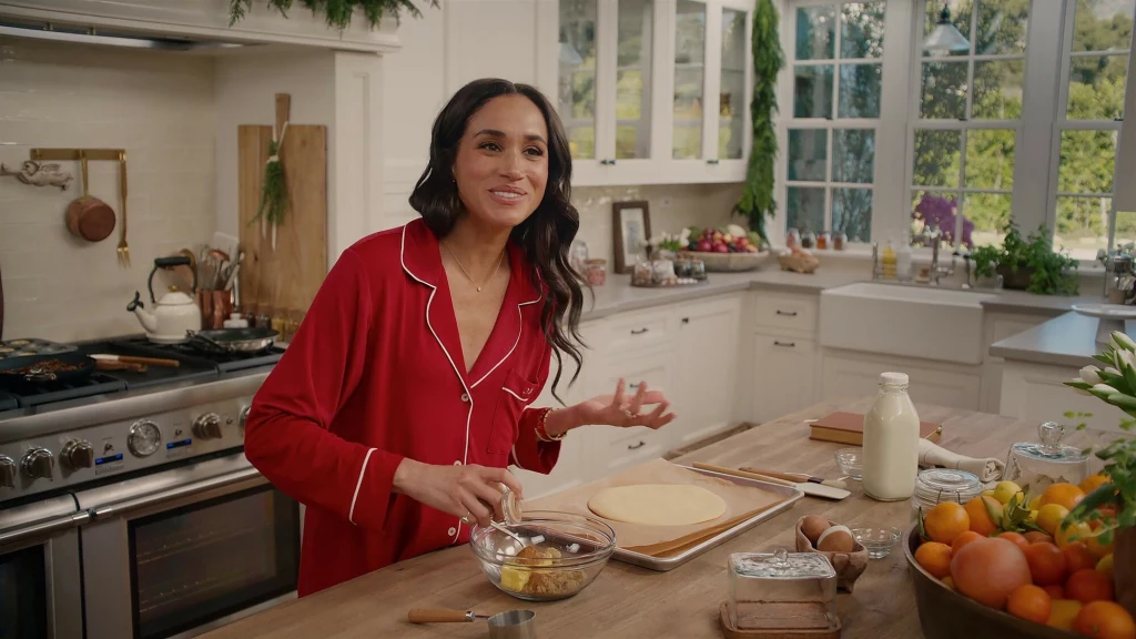 Meghan Markle zaskoczyła świątecznym przepisem. Jada się to u niej w domu tylko raz w roku Kobieta w czerwonej piżamie przygotowuje ciasto w jasnej, nowoczesnej kuchni. Przed nią na blacie leżą składniki takie jak jajka, mleko, świeże owoce oraz miska z masłem.
