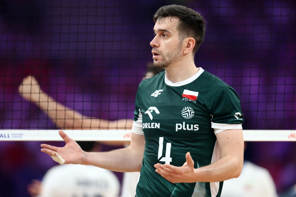 Siatkarz w zielonym stroju z orzełkiem i polską flagą na piersi stoi na boisku, gestykuluje rękami jakby okazywał zaskoczenie lub niezadowolenie, w tle inni gracze i siatka.