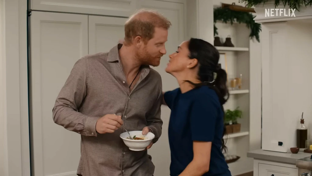 Książę Harry pokochał gumbo przygotowywane przez teściową Para młodych ludzi w przytulnej kuchni, mężczyzna trzyma w dłoni miskę z jedzeniem, uśmiechnięta kobieta pochyla się w kierunku niego jakby chciała go pocałować. Na tle jasne szafki i ozdoby świąteczne.