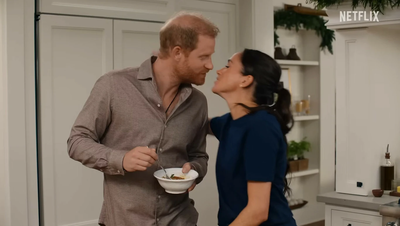 Para młodych ludzi w przytulnej kuchni, mężczyzna trzyma w dłoni miskę z jedzeniem, uśmiechnięta kobieta pochyla się w kierunku niego jakby chciała go pocałować. Na tle jasne szafki i ozdoby świąteczne.