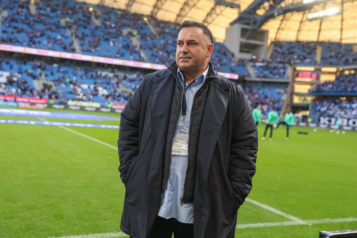 Mężczyzna w ciemnym płaszczu stoi na murawie stadionu piłkarskiego, za nim widoczne trybuny z kibicami oraz elementy infrastruktury stadionowej.