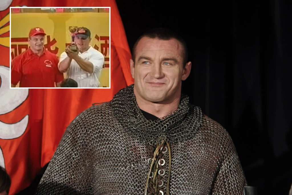 Mężczyzna w średniowiecznej kolczudze na tle czerwonej flagi z białym logo, wyrażający pewność siebie. W lewym górnym rogu wstawka przedstawiająca dwóch mężczyzn ubranych na czerwono, z jednym z nich trzymającym mikrofon lub rekwizyt.