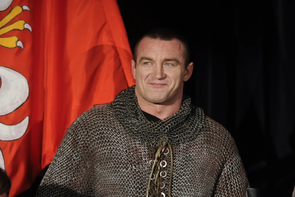 Mężczyzna w kolczudze z uśmiechem na twarzy, stojący na tle czerwonej flagi z heraldycznym symbolem.