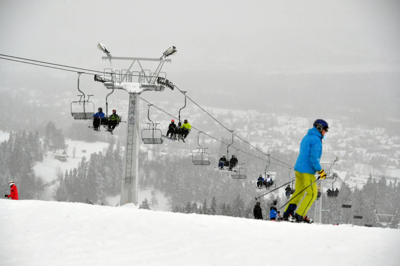Narciarz w niebieskiej kurtce i żółtych spodniach zjeżdża po ośnieżonym stoku narciarskim, w tle widać wyciąg krzesełkowy z osobami jadącymi na górę oraz zabudowania miasta pokryte śniegiem.