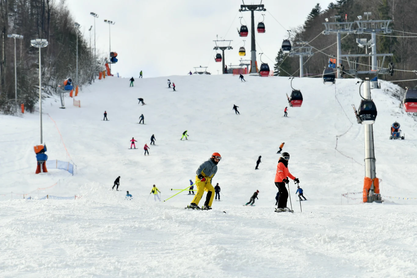 Coraz więcej ośrodków narciarskich otwiera stoki Narciarze zjeżdżający po szerokim, ośnieżonym stoku, nad którym unoszą się gondole i wyciągi narciarskie. W tle liczni ludzie korzystający z atrakcji zimowych. Wokół stoku rośnie las iglasty.