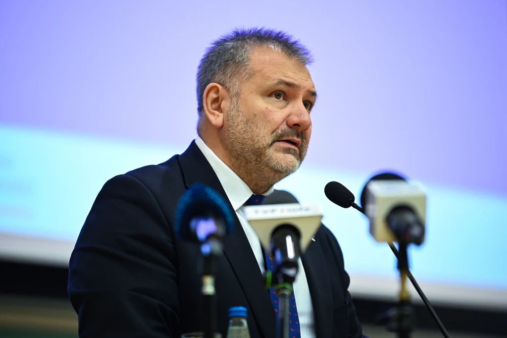 Mężczyzna w garniturze przemawia do mikrofonów podczas konferencji lub spotkania, w tle widoczny rozmyty ekran prezentacji.