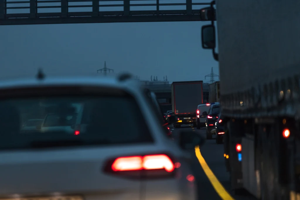 W jakich warunkach należy włączyć światła przeciwmgłowe? Korek na autostradzie w godzinach wieczornych, widoczne samochody i ciężarówki ustawione jeden za drugim na wielopasmowej drodze, światła pojazdów podkreślają atmosferę zmierzchu.