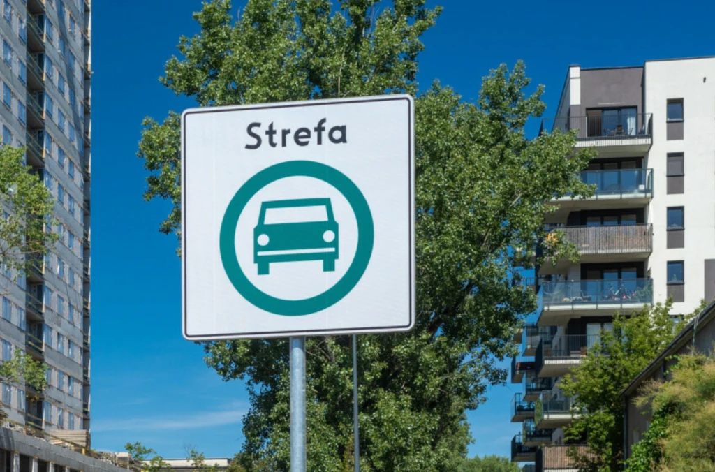 Znak drogowy z napisem „Strefa” oraz symbolem samochodu na tle nowoczesnych budynków mieszkalnych i niebieskiego nieba.