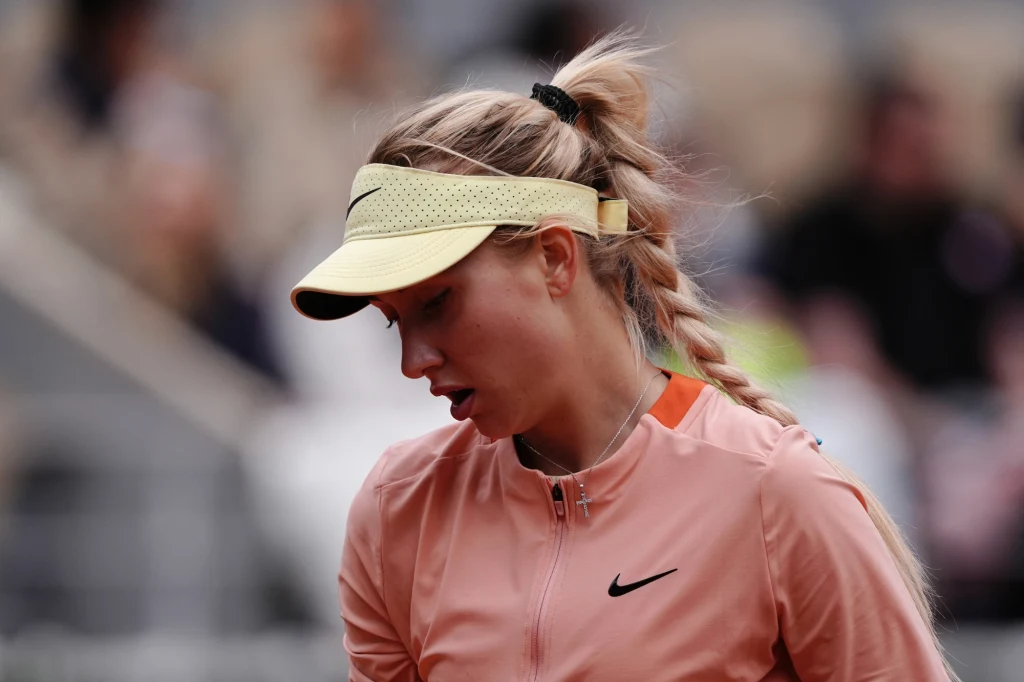 Anastazja Potapowa w trakcie "lania" od Igi Świątek. Paryż 2024 Młoda kobieta z długim blond warkoczem i sportową czapeczką z daszkiem, ubrana w różową sportową bluzę z widocznym logo Nike, pochylona głową na tle rozmytego stadionu.