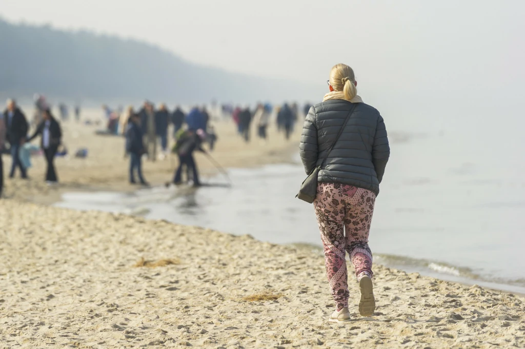 Kobieta ubrana w ciemną kurtkę i wzorzyste spodnie spaceruje brzegiem plaży, w tle liczna grupa ludzi rozproszonych wzdłuż wybrzeża, zamglone niebo nad morzem oraz las tworzą spokojną atmosferę.
