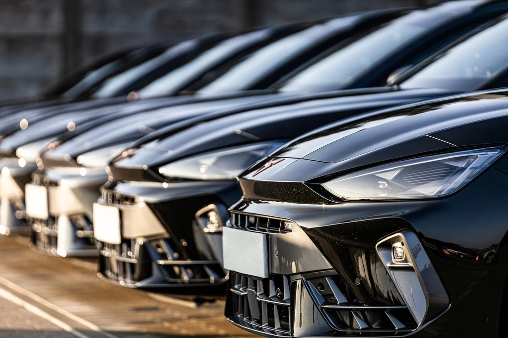 Rząd nowoczesnych czarnych samochodów ustawionych w jednej linii na parkingu, widoczne odbicia światła na błyszczących maskach pojazdów, jednolity, dynamiczny charakter kompozycji, skoncentrowany na detalach przodu aut.