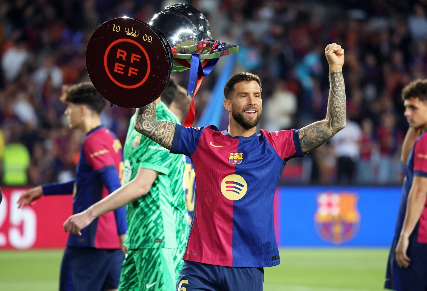 Piłkarz drużyny FC Barcelona w stroju meczowym trzyma w górze trofeum, świętując zwycięstwo na stadionie w otoczeniu innych zawodników.