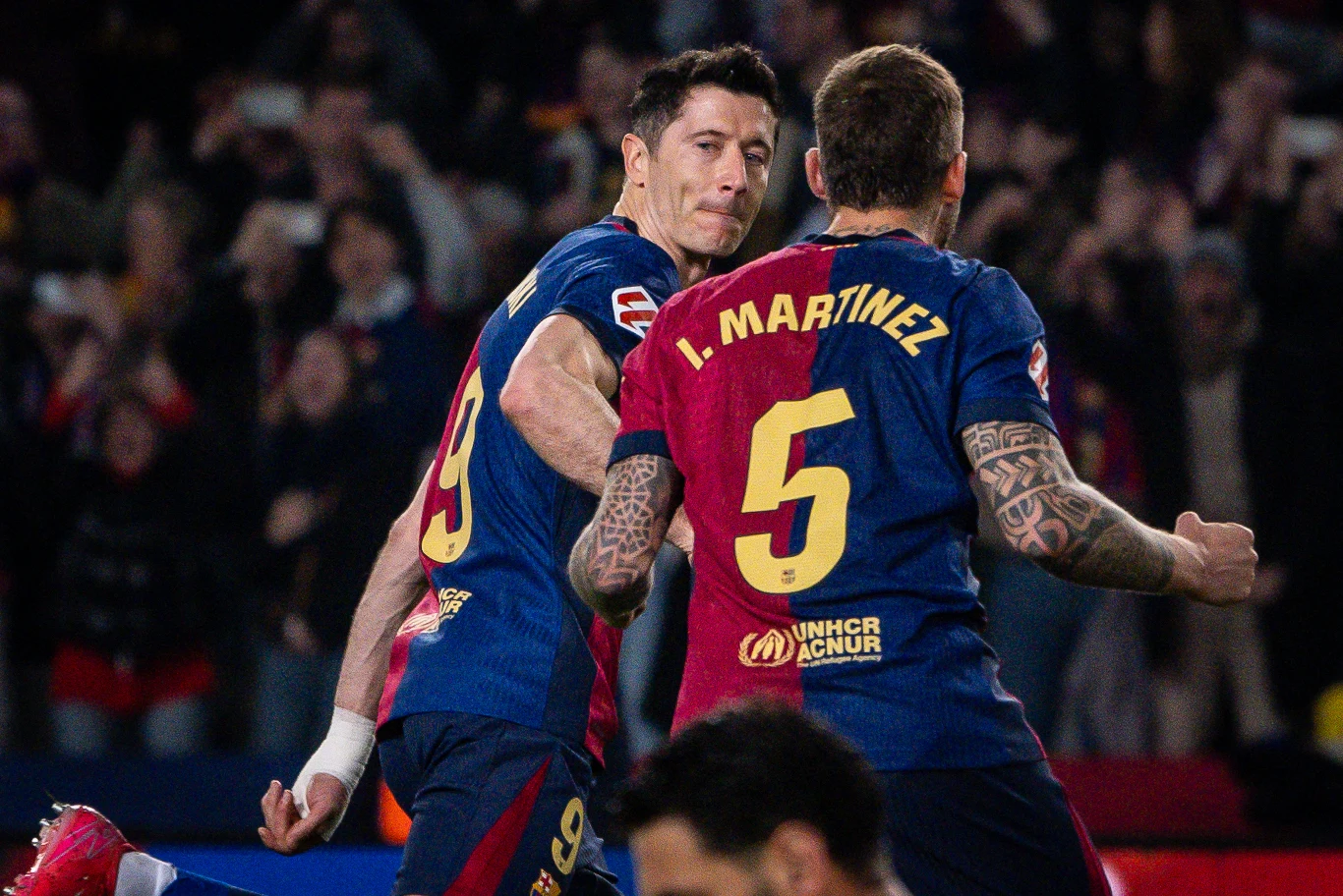 Dwóch piłkarzy w granatowo-bordowych strojach na tle tłumu kibiców podczas meczu, jeden z zawodników z numerem 9 unosi rękę w geście triumfu, drugi ma widoczny tatuaż na ręce i numer 5 na plecach.