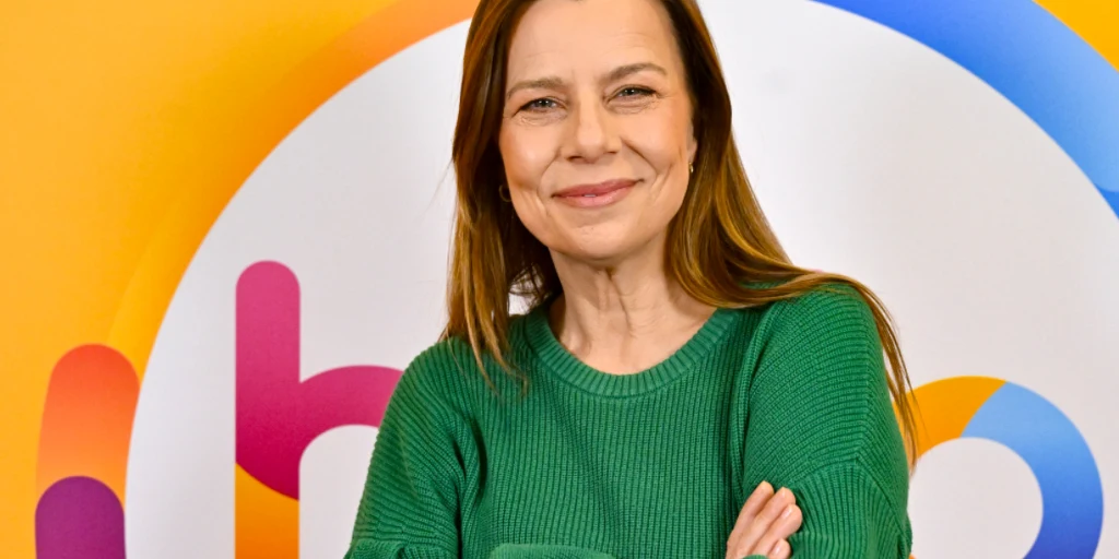 Agata Kulesza Kobieta w zielonym swetrze z długimi, brązowymi włosami uśmiecha się, stojąc na tle kolorowego logo z okręgiem.