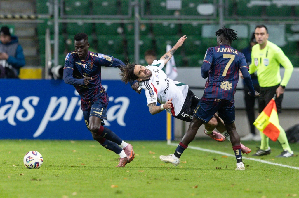 Dwóch piłkarzy w niebieskich strojach walczy o piłkę z zawodnikiem w białym stroju, który upada na murawę boiska, w tle sędziowie oraz trybuny.