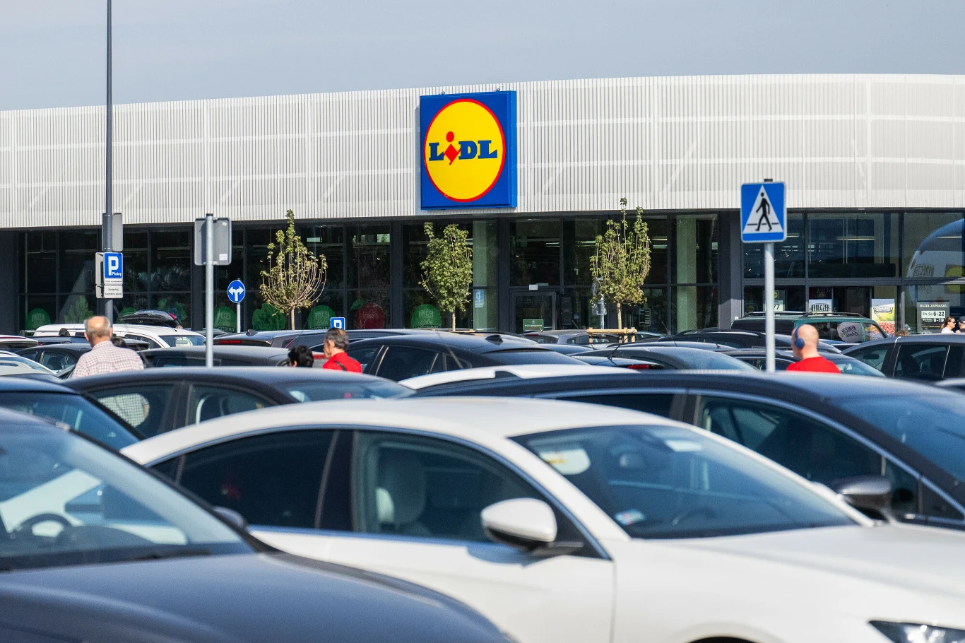 Samochody zaparkowane na dużym parkingu przed supermarketem Lidl, w tle widoczne wejście do sklepu oznaczone charakterystycznym logo.
