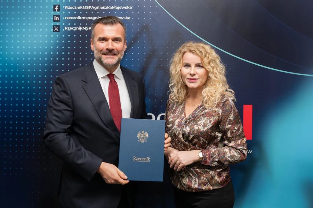 Dwie osoby w stroju biznesowym stoją obok siebie na tle z elementami graficznymi oraz nazwą instytucji. Mężczyzna trzyma w ręku teczkę z godłem państwowym i napisem Rzecznik.