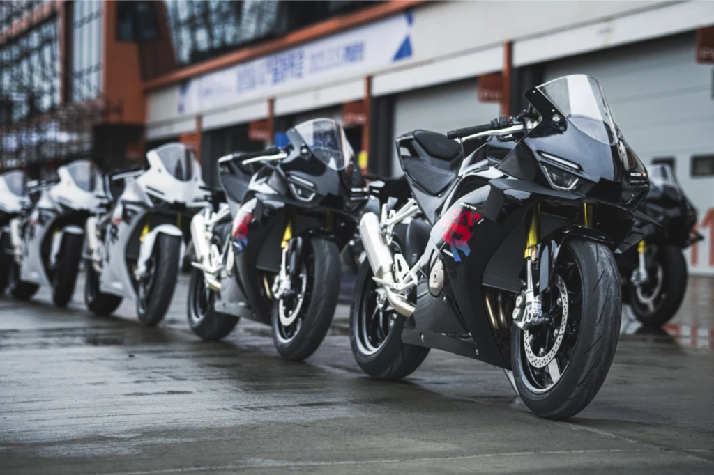 Kolejna chińska marka motocykli zadebiutuje w Europie. Rząd nowoczesnych, sportowych motocykli ustawionych na mokrym asfalcie przed budynkami garażowymi toru wyścigowego, każdy z nich posiada aerodynamiczną osłonę i charakterystyczne kolorowe oznaczenia.