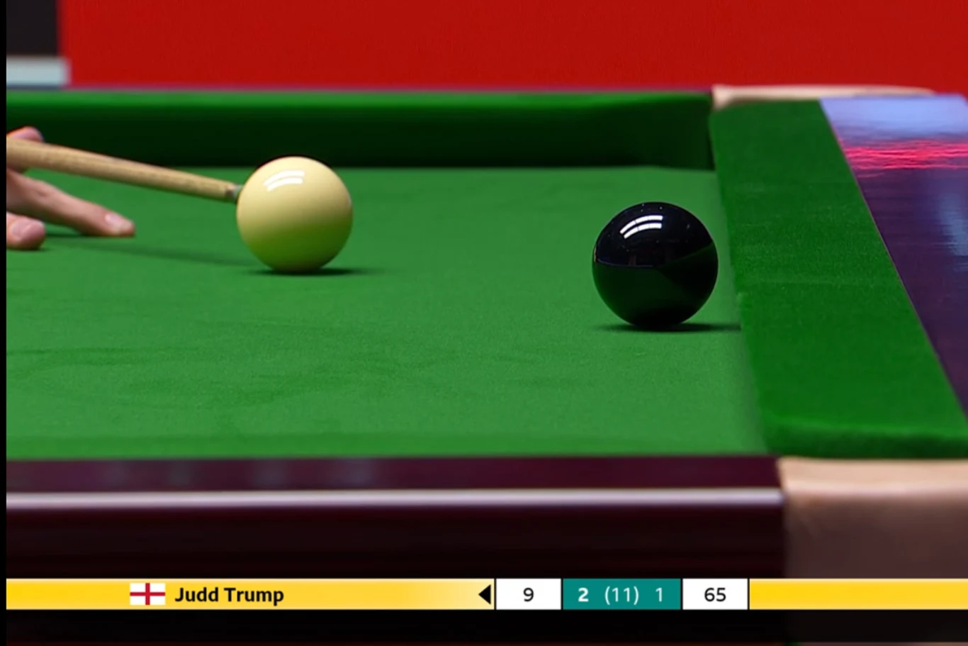 Stół bilardowy z zielonym suknem, żółta bila przygotowana do uderzenia przez gracza oraz czarna bila ustawiona na stole. W tle wycinek dłoni trzymającej kij, na dole pasek informacyjny z nazwiskiem zawodnika i wynikiem meczu.