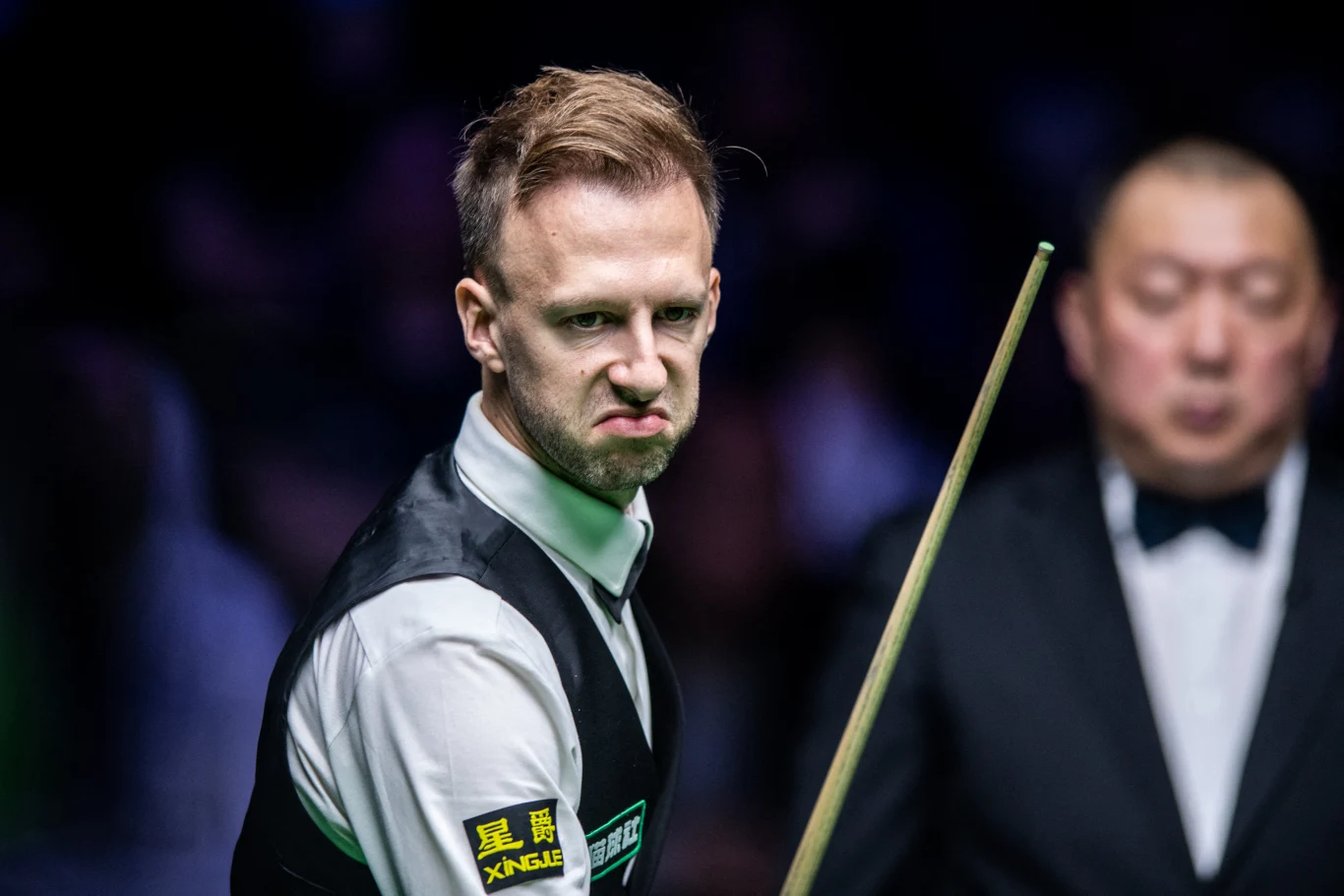 Zawodnik snookera w kamizelce i koszuli z wyraźnie zaciętą miną, trzymający kij bilardowy, w tle widoczny sędzia ubranego w ciemny garnitur z muchą.