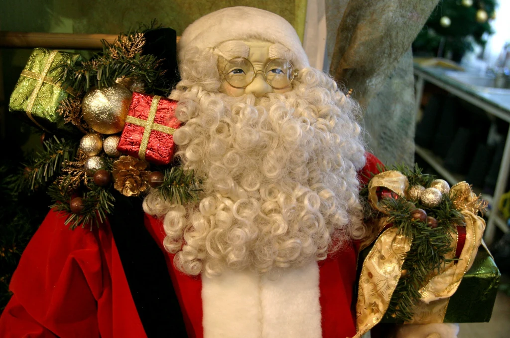 Figurka Mikołaja z siwą brodą i okularami, ubrana w czerwony strój, trzymająca ozdobione prezentami i złotymi dekoracjami gałązki świerkowe.