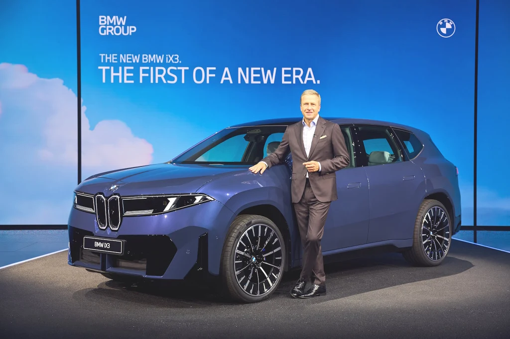 Mężczyzna w garniturze opiera się o niebieski samochód marki BMW IX3 na scenie podczas oficjalnej prezentacji, w tle widoczna duża grafika z napisem 'The first of a new era'.