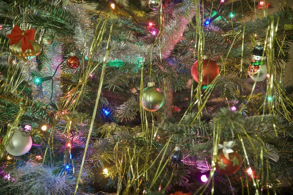 Ozdoby choinkowe w postaci kolorowych bombek, srebrnych i złotych łańcuchów oraz lampek tworzących świąteczną iluminację na zielonych gałęziach choinki.