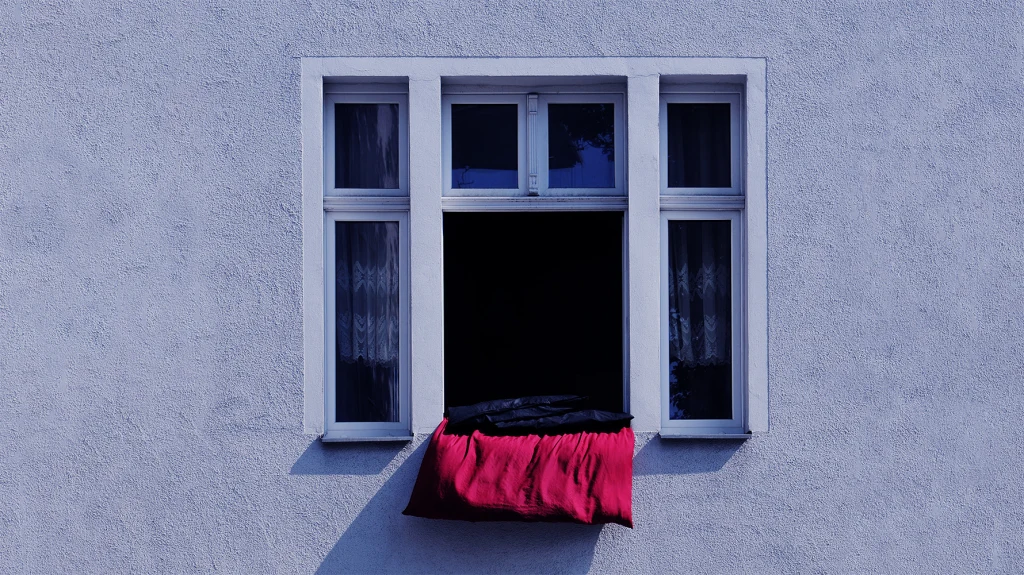 Otwór w białej tynkowanej ścianie z oknem, przez które przewieszony jest czerwony koc lub duży materiał, a wnętrze pomieszczenia pozostaje zaciemnione