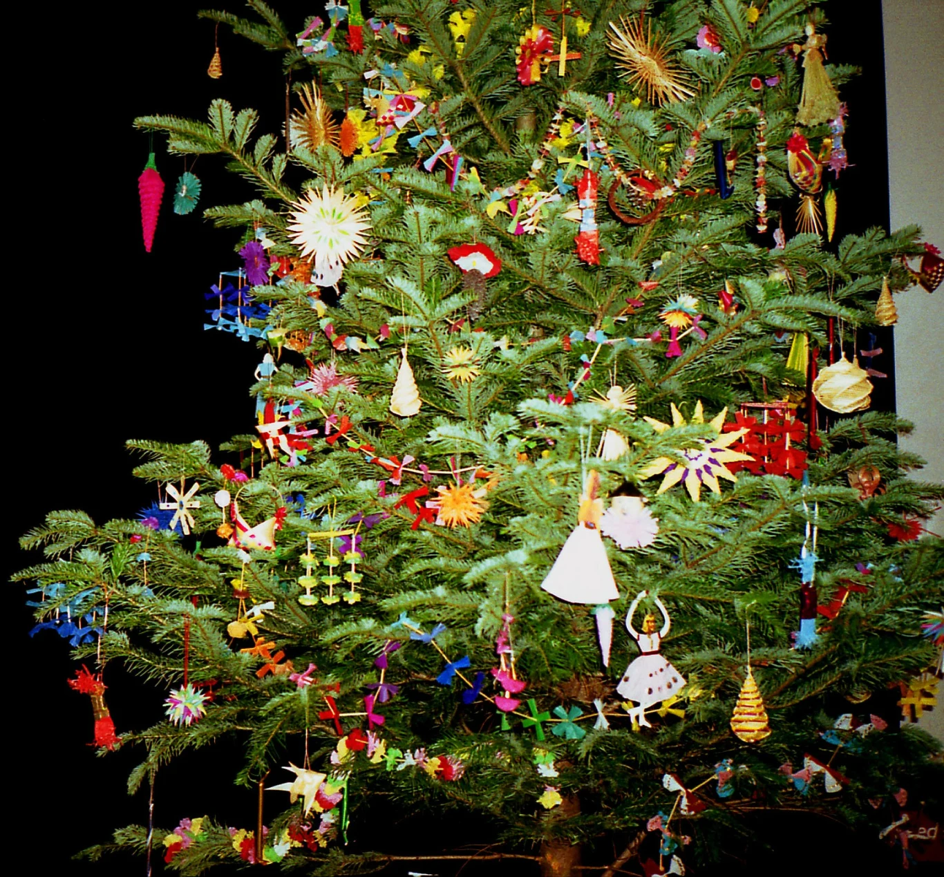 Okazała choinka udekorowana wieloma kolorowymi ozdobami świątecznymi wykonanymi z papieru i innych materiałów, wśród których dominują gwiazdy, aniołki, ptaszki oraz wiszące girlandy.