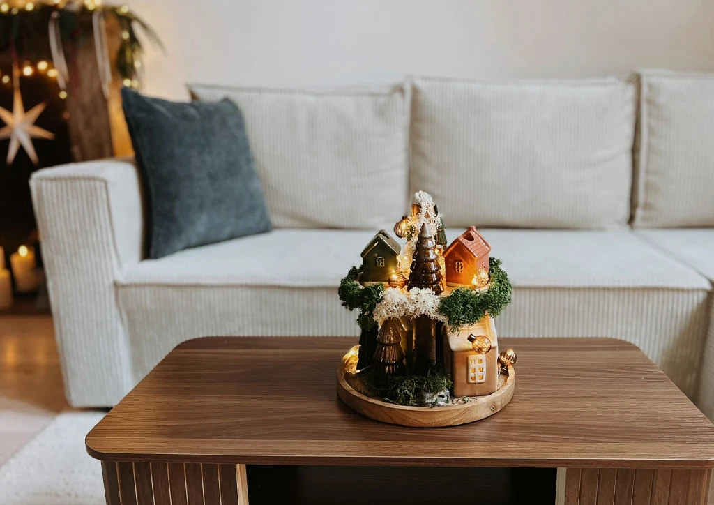 Dekoracja świąteczna w postaci miniaturowej wioski z domkami, drzewkiem i drobnymi światełkami ustawiona na drewnianym stoliku, w tle jasna sofa i przytulne wnętrze salonu.