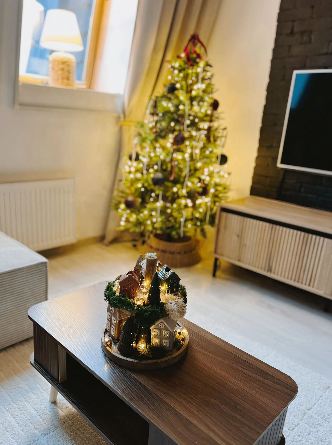 Wnętrze salonu z jasnymi ścianami, ustawioną po lewej stronie lampą w oknie, udekorowaną i oświetloną choinką w tle oraz nowoczesnymi meblami. Na pierwszym planie na drewnianym stole znajduje się bożonarodzeniowa dekoracja w postaci miniaturowego domku...