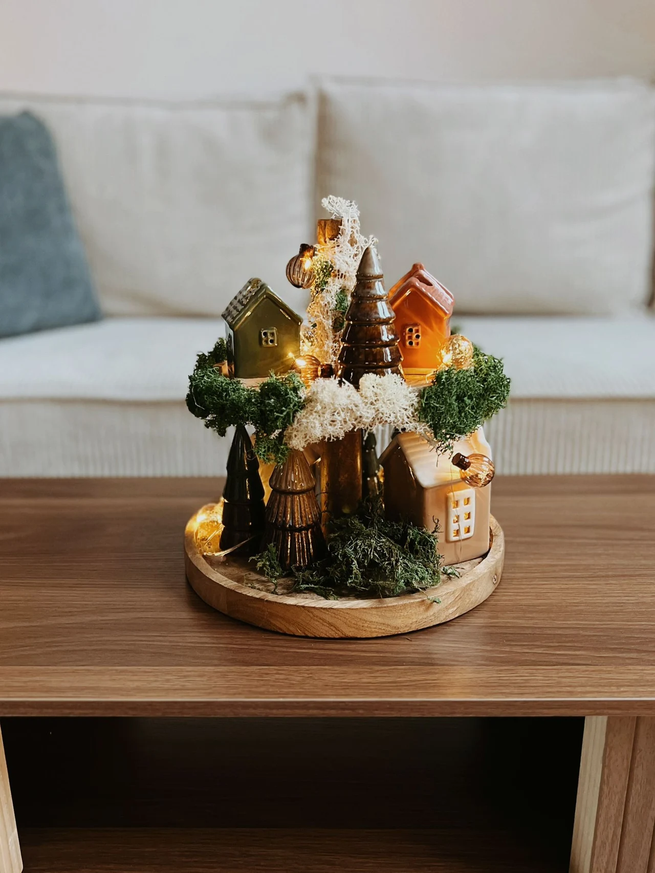 Miniaturowa dekoracja przedstawiająca małe, kolorowe domki i ozdobne drzewka ustawione na drewnianej podstawce, podświetlona drobnymi lampkami LED, stoi na drewnianym stoliku w nowoczesnym salonie.