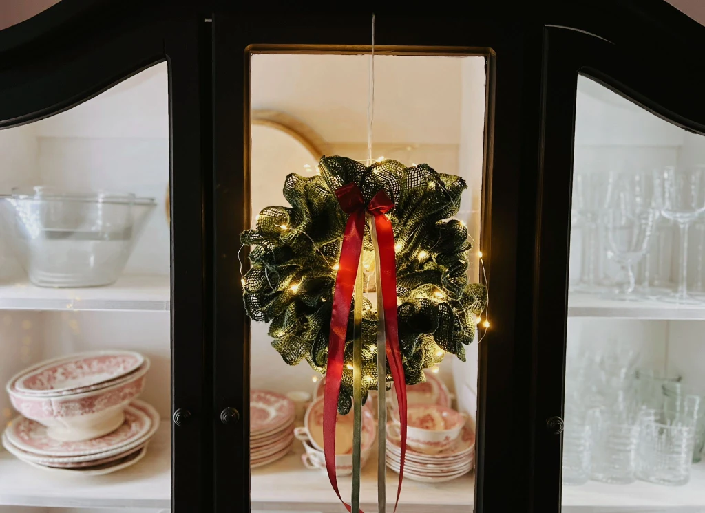 Zielony wieniec bożonarodzeniowy ozdobiony światełkami i czerwoną wstążką zawieszony na przeszklonych drzwiach kredensu. W tle porcelanowe miski, talerze i szklanki ustawione na półkach.