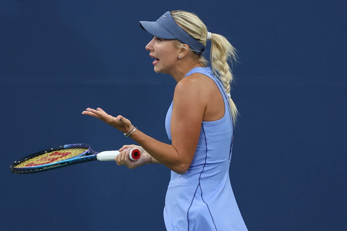 Anastazja Potapowa Kobieta w sportowej sukience i czapce z daszkiem podczas meczu tenisowego trzyma rakietę w jednej dłoni, drugą dłonią wykonuje gest wyrażający frustrację. Tło jednolicie granatowe, skupienie na emocjach zawodniczki.