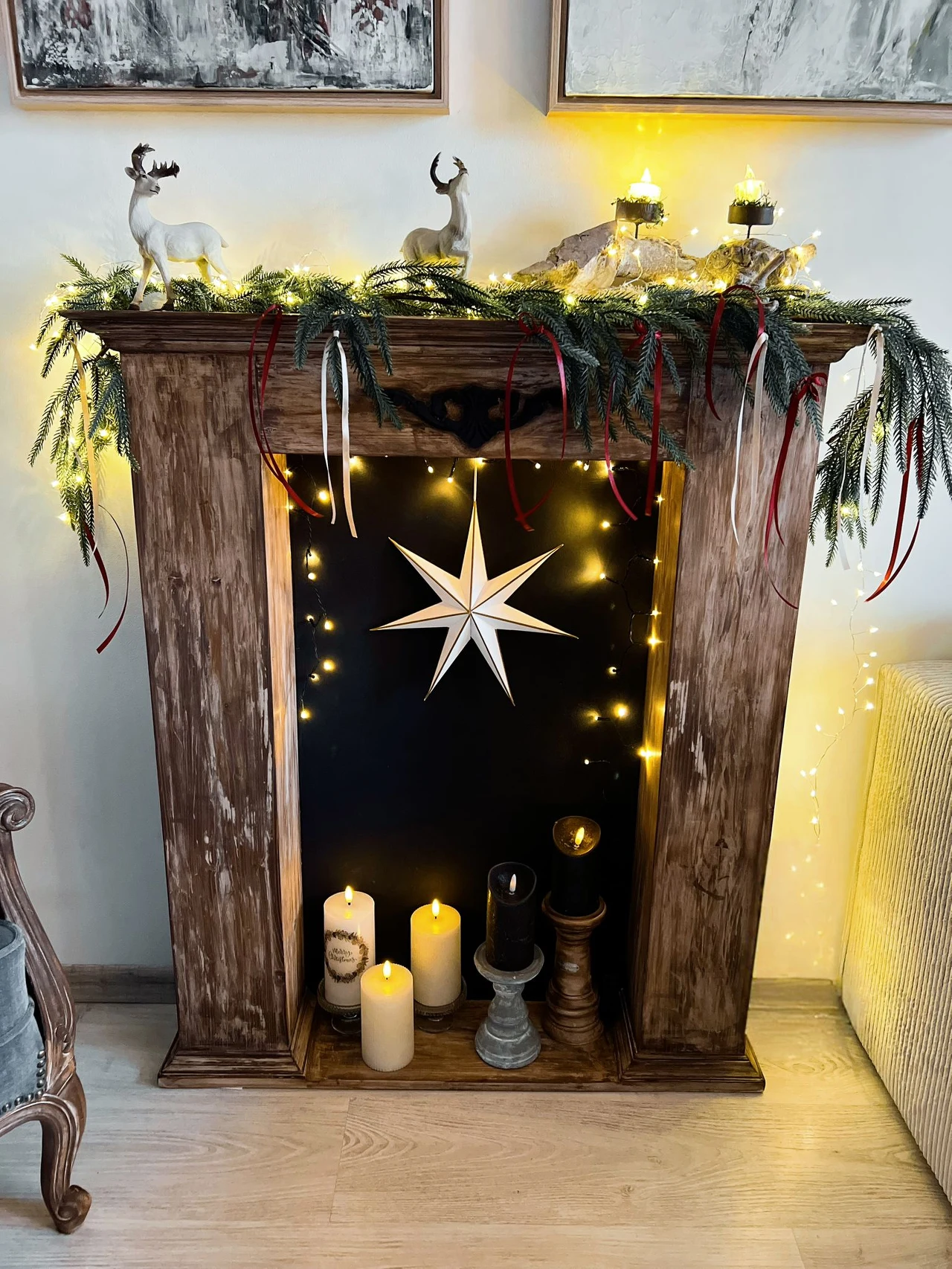 Drewniany portal kominkowy udekorowany świątecznym stroikiem z gałązkami, szyszkami i ozdobami reniferów, wewnątrz ustawione sztuczne świece i biała gwiazda na czarnym tle oraz światełka LED.